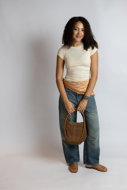 A woman stands against a light gray studio background wearing a cream short-sleeve top, blue jeans, brown woven shoes, and holding a brown leather shoulder bag.