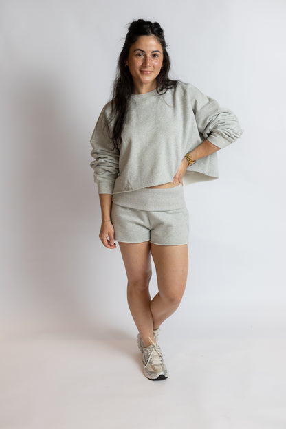 A woman modeling a light gray heathered lounge set consisting of a cropped long-sleeve sweatshirt and matching shorts, paired with gray sneakers, against a plain light gray studio background.