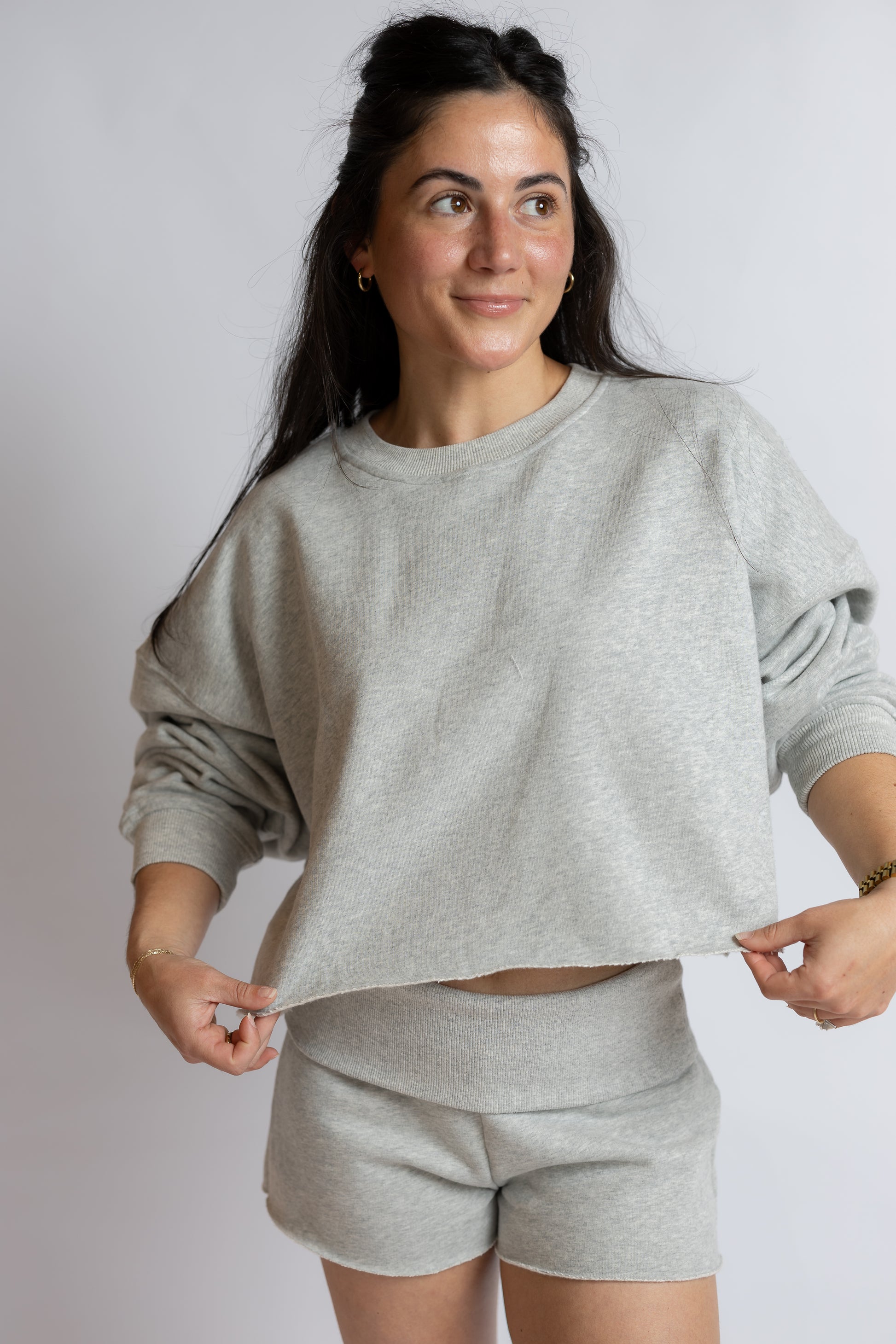 A woman modeling a light gray heathered crewneck sweatshirt and matching shorts against a plain light gray studio background.