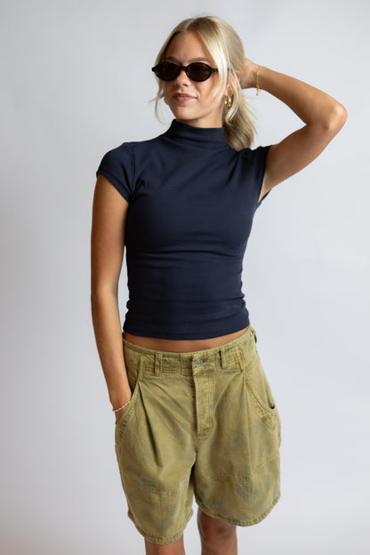 A woman modeling a dark navy blue mock neck short-sleeve crop top paired with olive green cargo shorts, set against a plain light gray studio background.