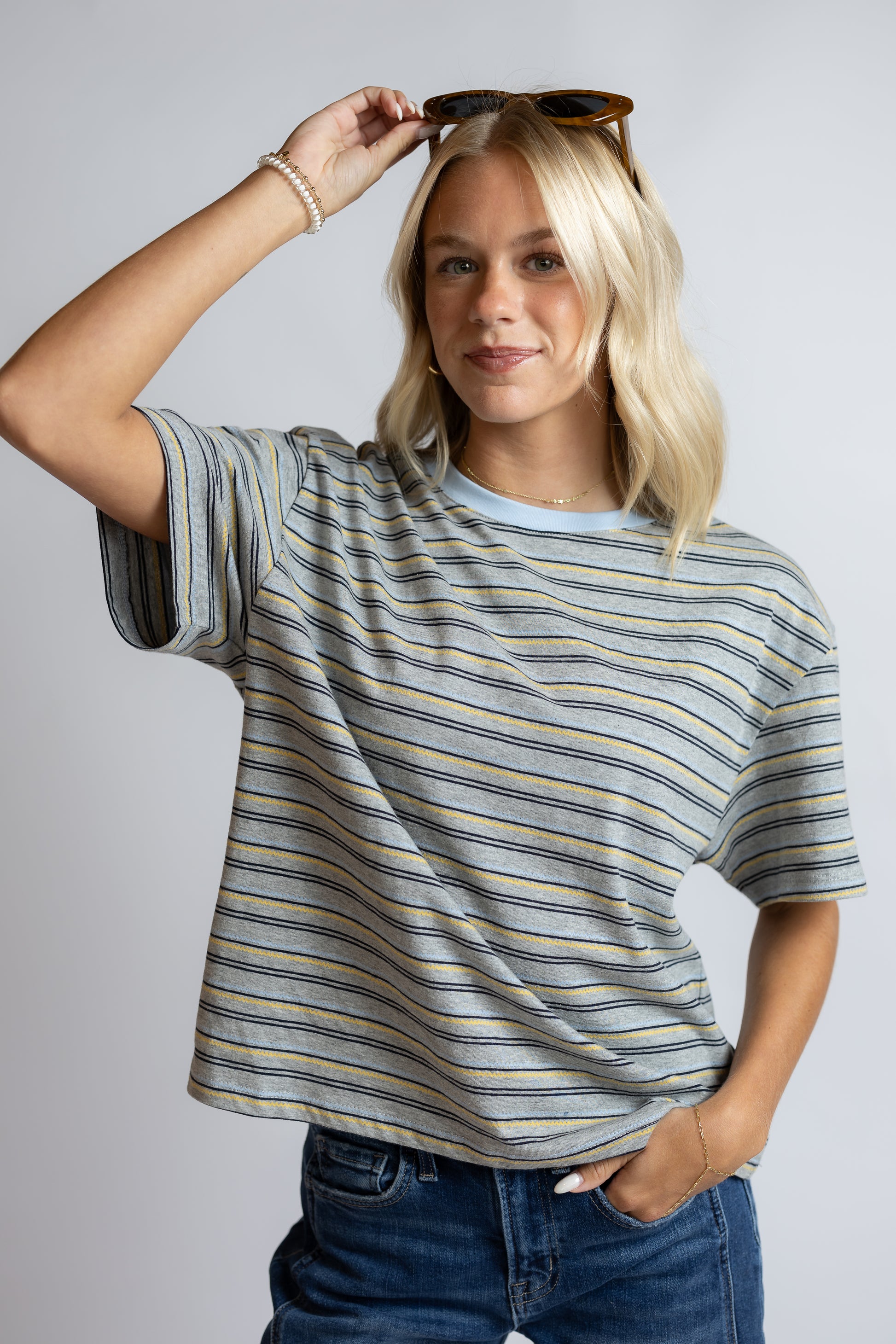 A woman modeling a short-sleeve crewneck t-shirt with thin horizontal stripes in blue, yellow, and gray, paired with blue jeans against a light gray studio background.