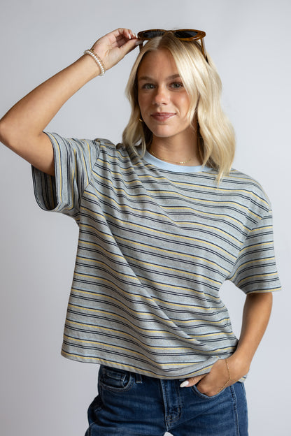 A woman modeling a short-sleeve crewneck t-shirt with thin horizontal stripes in blue, yellow, and gray, paired with blue jeans against a light gray studio background.