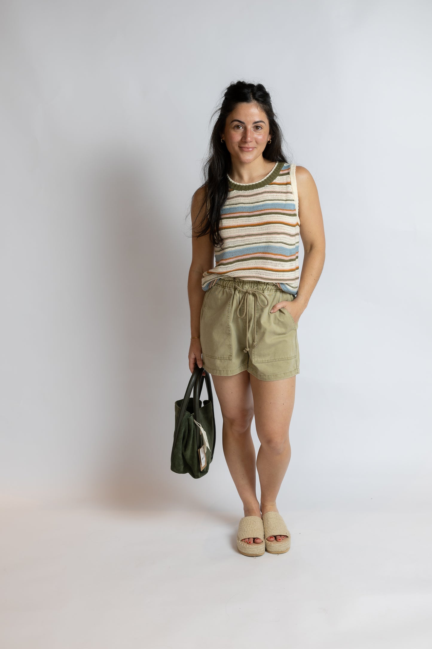 A woman stands against a light gray studio background wearing a sleeveless striped tank top with green trim, olive green drawstring shorts, beige slide sandals, and holding a dark green tote bag.