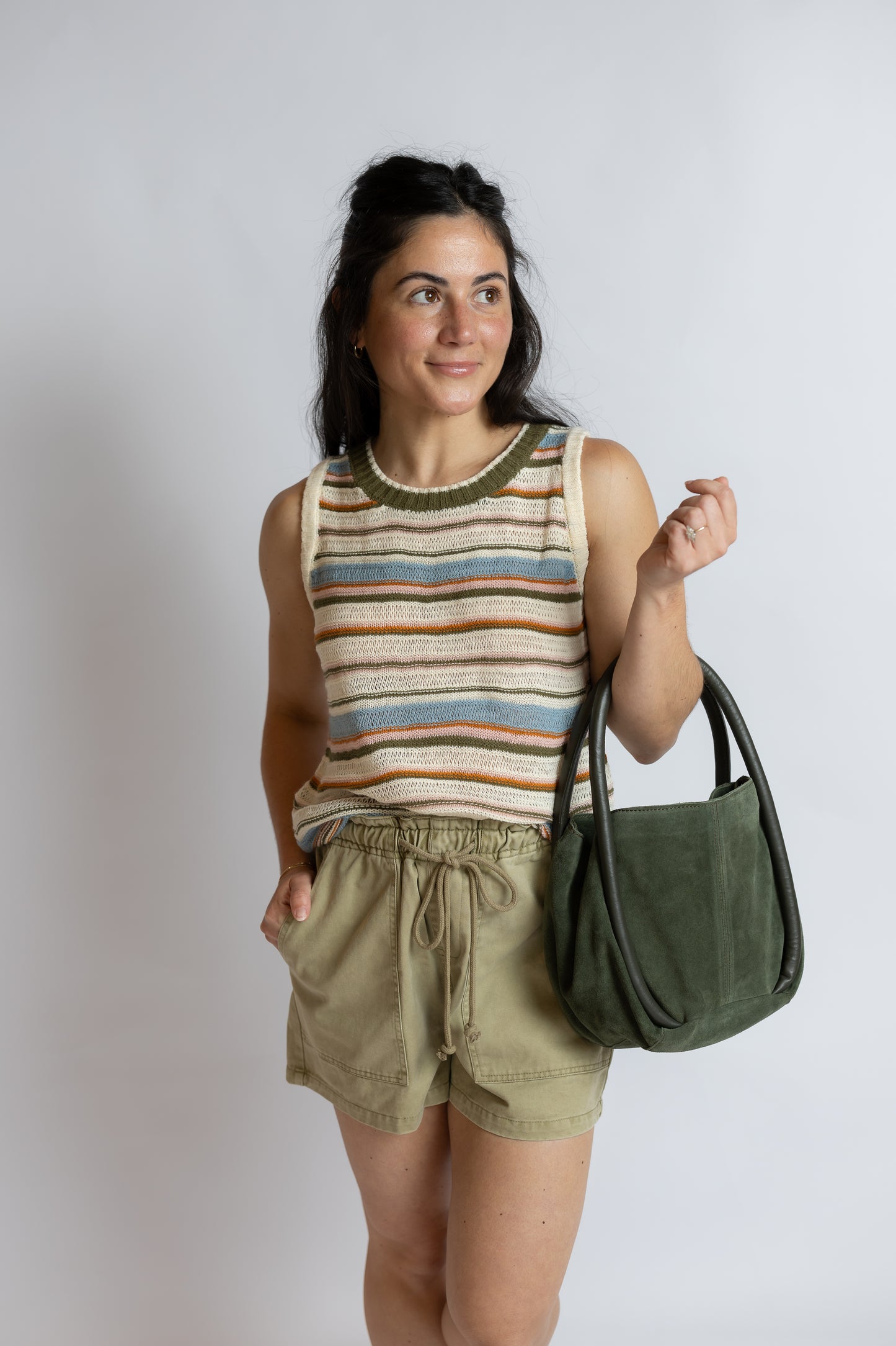 A woman modeling a striped knit tank top, olive green drawstring shorts, and carrying an olive green suede shoulder bag against a light gray studio background.