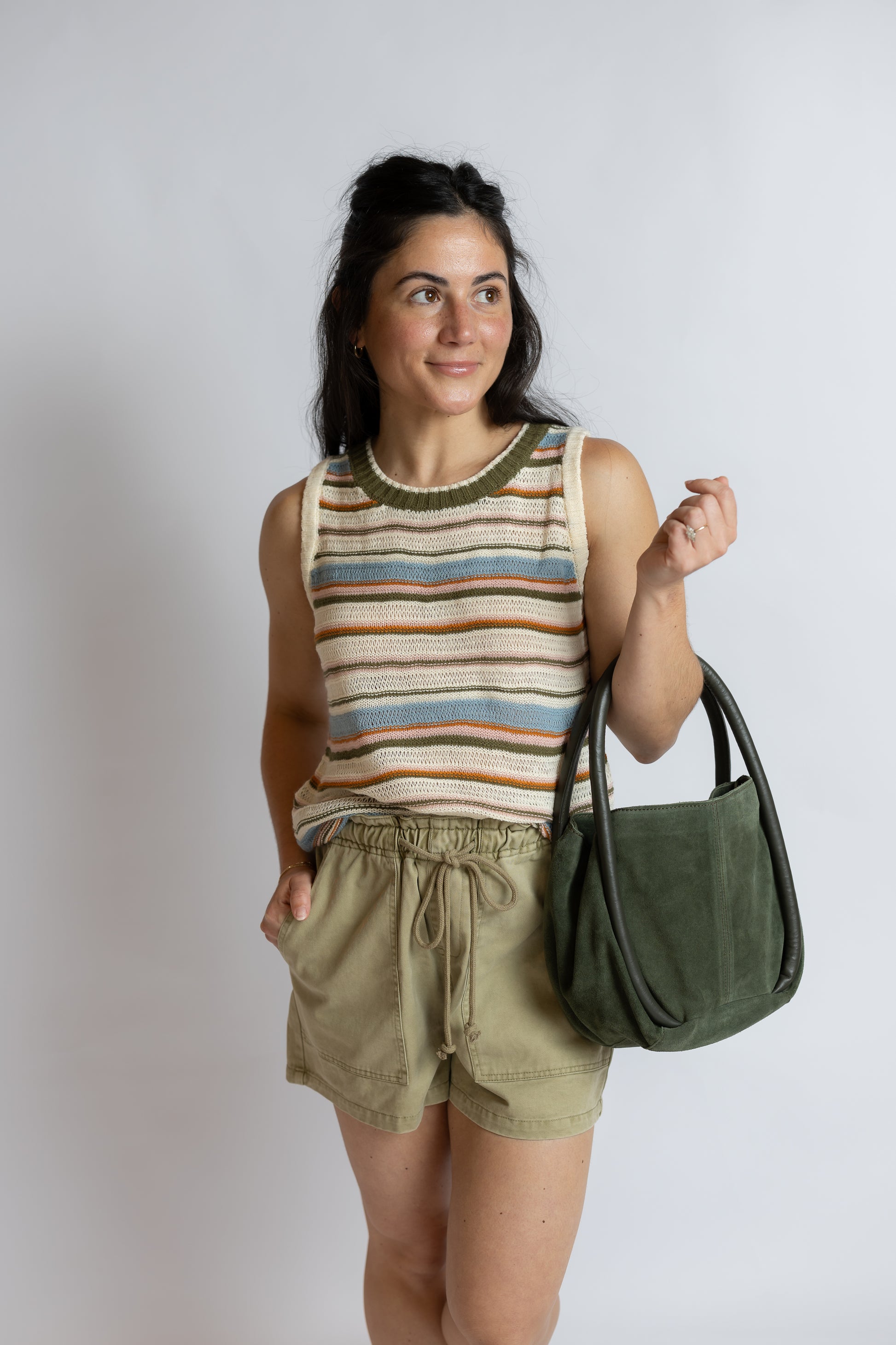 A woman modeling a striped knit tank top, olive green drawstring shorts, and carrying an olive green suede shoulder bag against a light gray studio background.