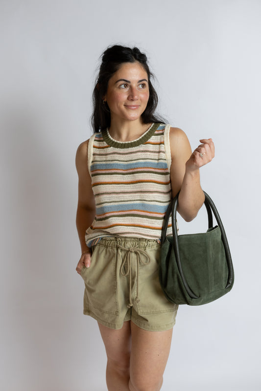 A woman modeling a striped knit tank top, olive green drawstring shorts, and carrying an olive green suede shoulder bag against a light gray studio background.