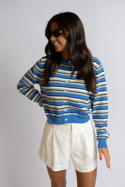 A woman wearing a blue, white, and beige striped long-sleeve knit cardigan with white shorts, posing against a light gray studio background.