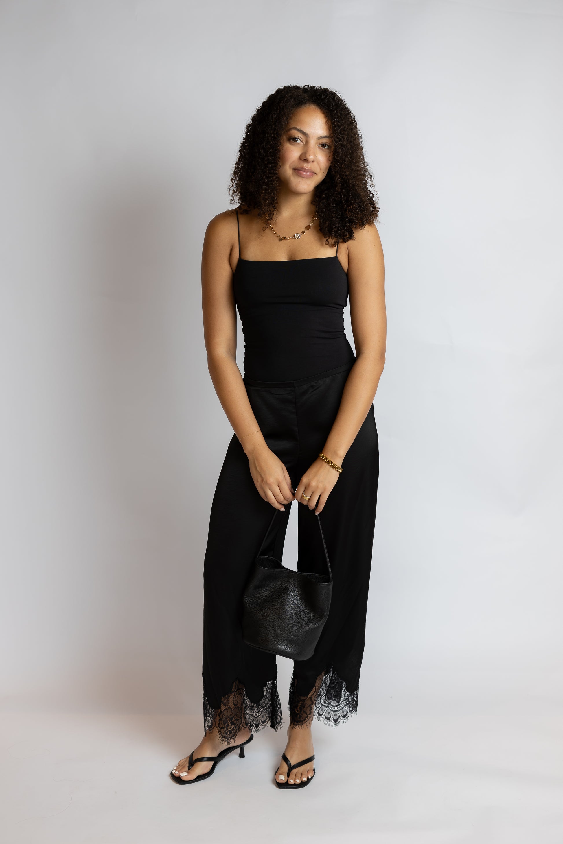 A woman modeling a black spaghetti-strap jumpsuit with black lace trim at the hem, paired with black sandals and holding a black handbag, against a light gray studio background.