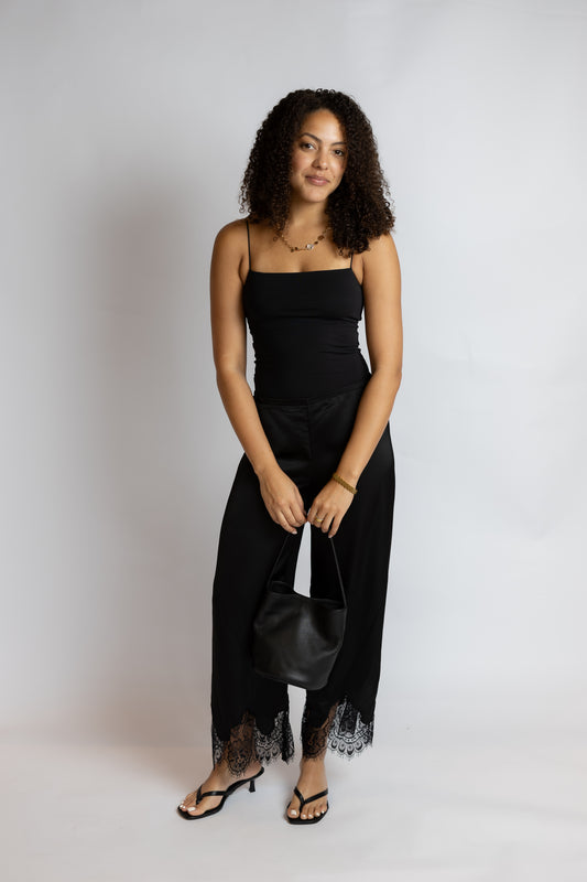 A woman modeling a black spaghetti-strap jumpsuit with black lace trim at the hem, paired with black sandals and holding a black handbag, against a light gray studio background.