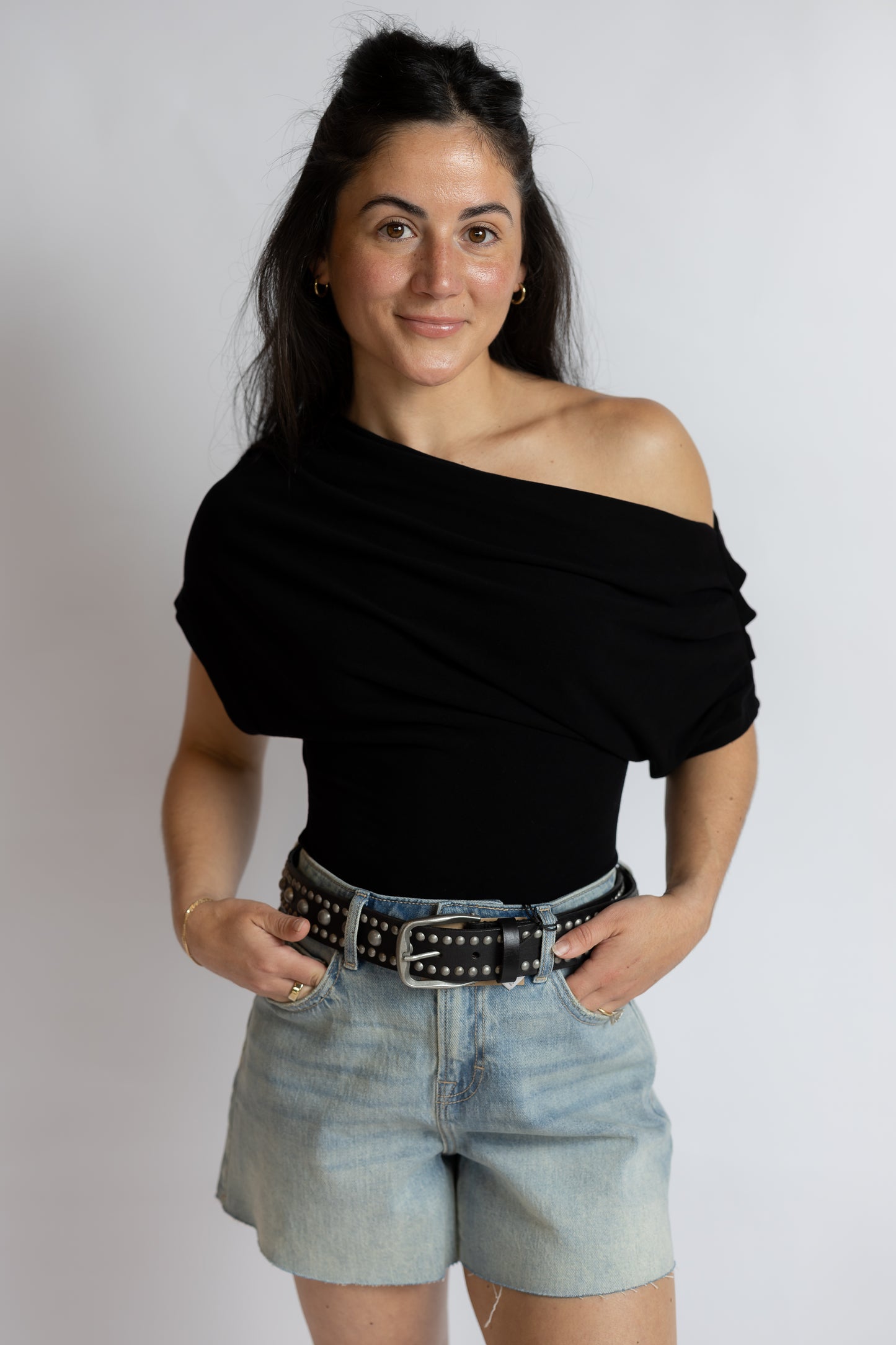 A woman modeling a black off-the-shoulder draped top paired with light-wash denim shorts and a black studded belt, standing against a plain light gray background.