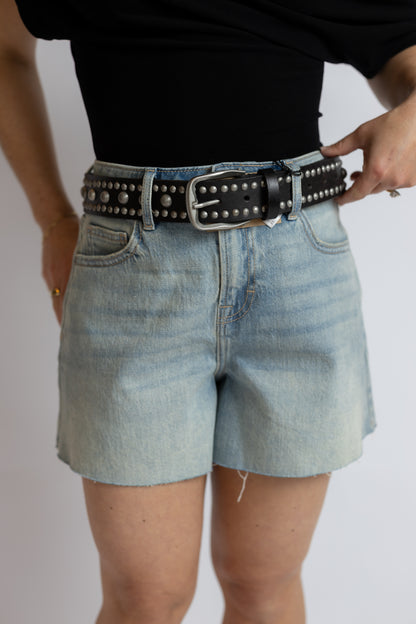 A person wearing light-wash denim shorts and a black studded belt with a silver buckle, shown from the chest to the upper thighs against a plain light background.