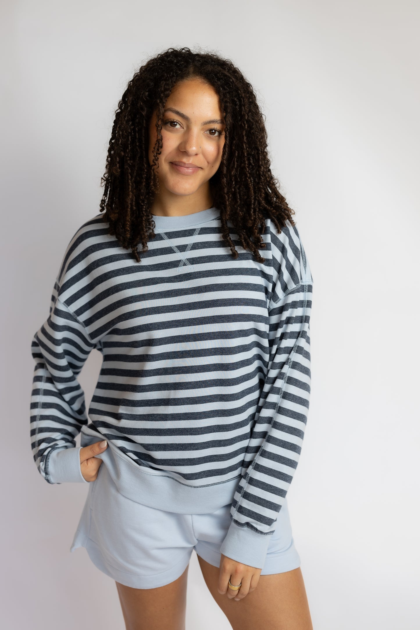 A woman modeling a long-sleeve crewneck sweatshirt with navy and light blue horizontal stripes, paired with light blue shorts, against a plain light gray background.