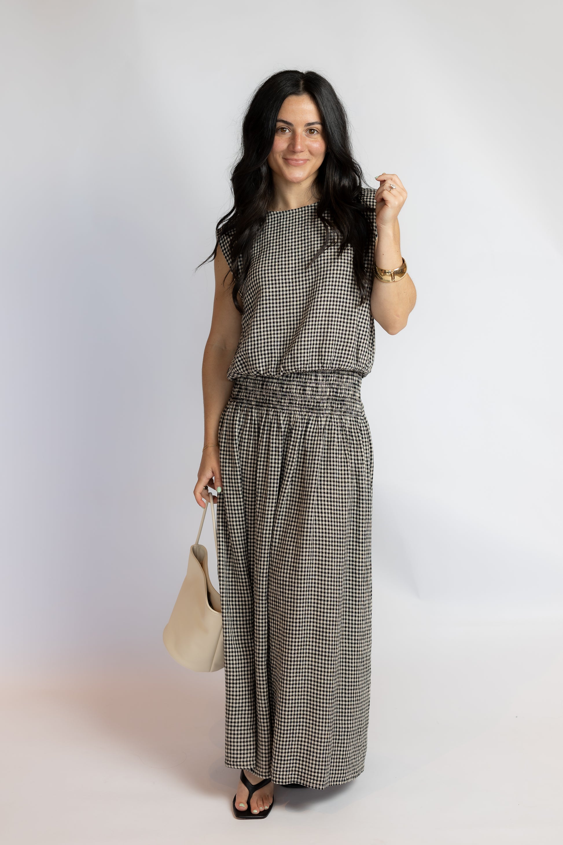 A woman modeling a sleeveless, wide-leg jumpsuit in a black and white gingham pattern, holding a beige tote bag against a plain light gray studio background.