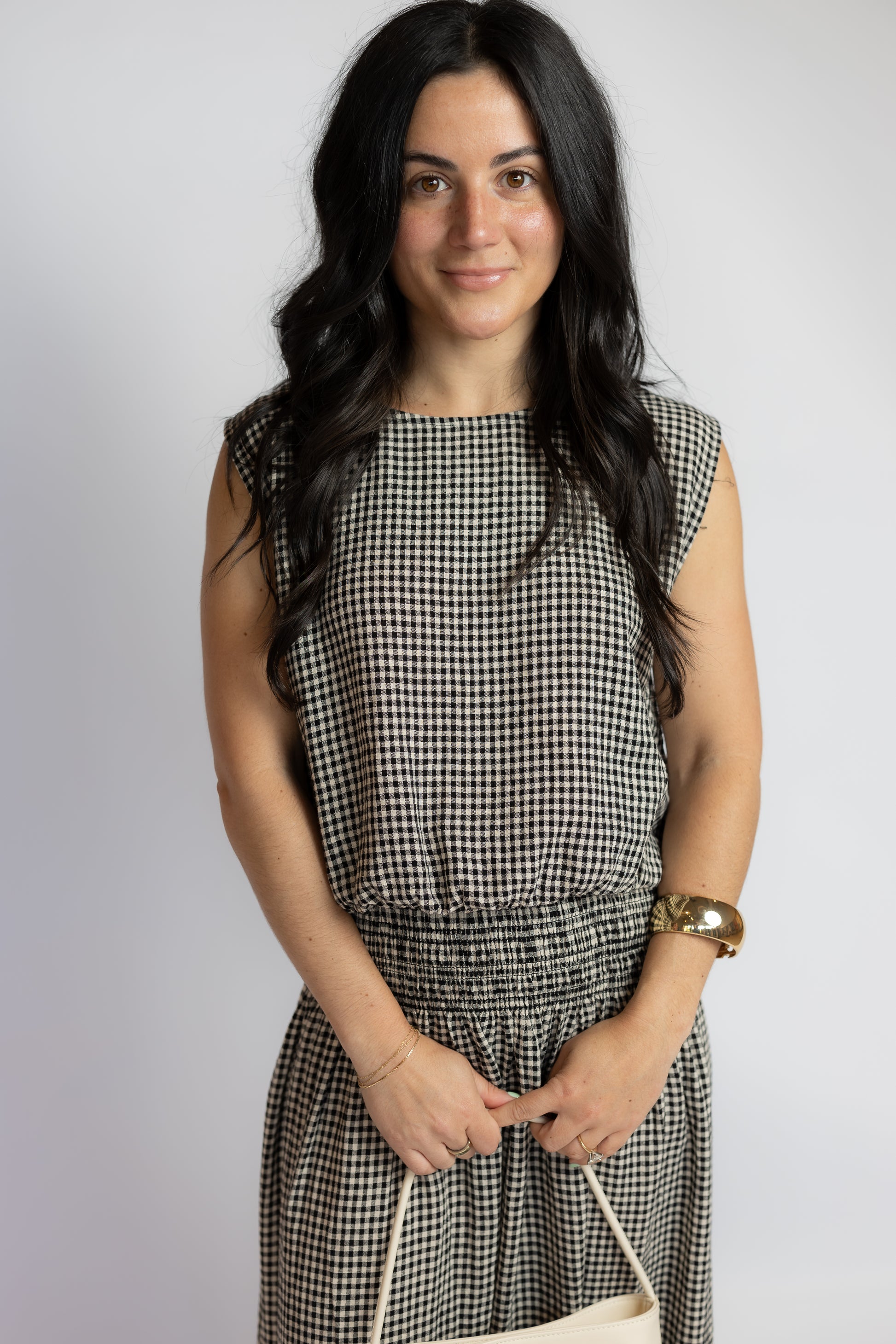 A woman with long dark hair wearing a sleeveless black and white gingham dress, holding a cream-colored handbag against a plain light gray background.