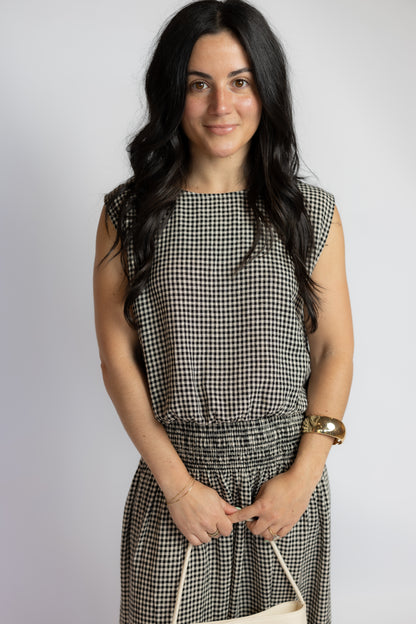 A woman with long dark hair wearing a sleeveless black and white gingham dress, holding a cream-colored handbag against a plain light gray background.