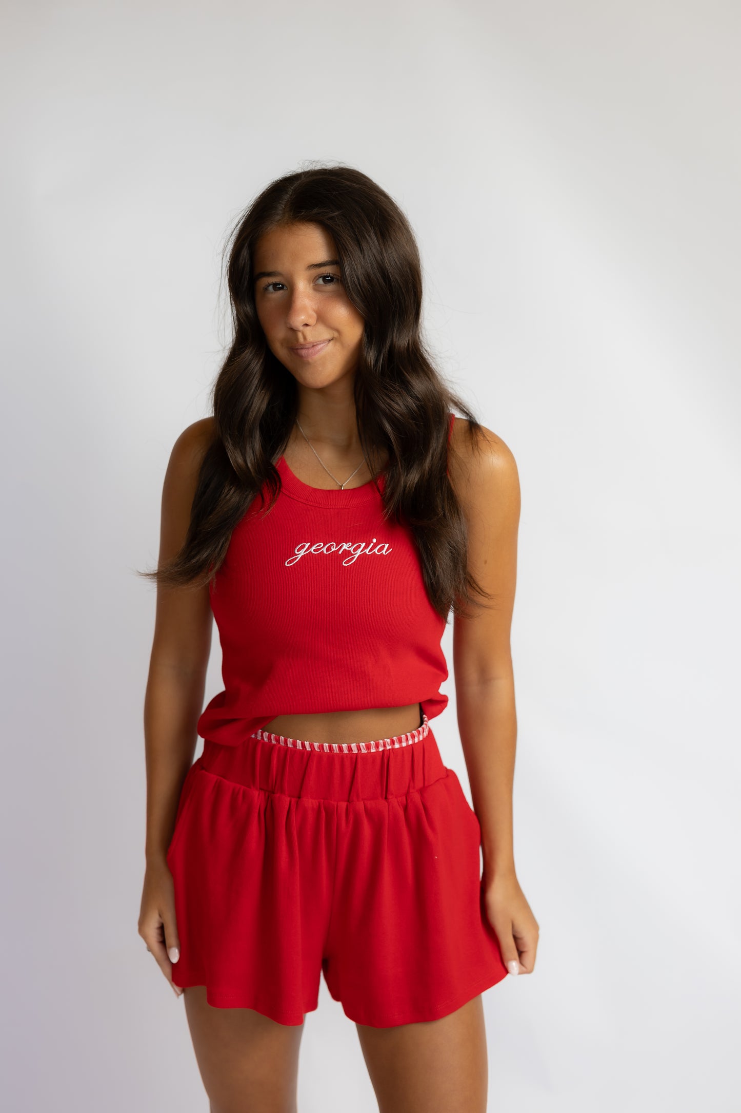 A woman modeling a red sleeveless crop top and matching red shorts against a light gray studio background. The top features the word 'georgia' in white script across the chest.