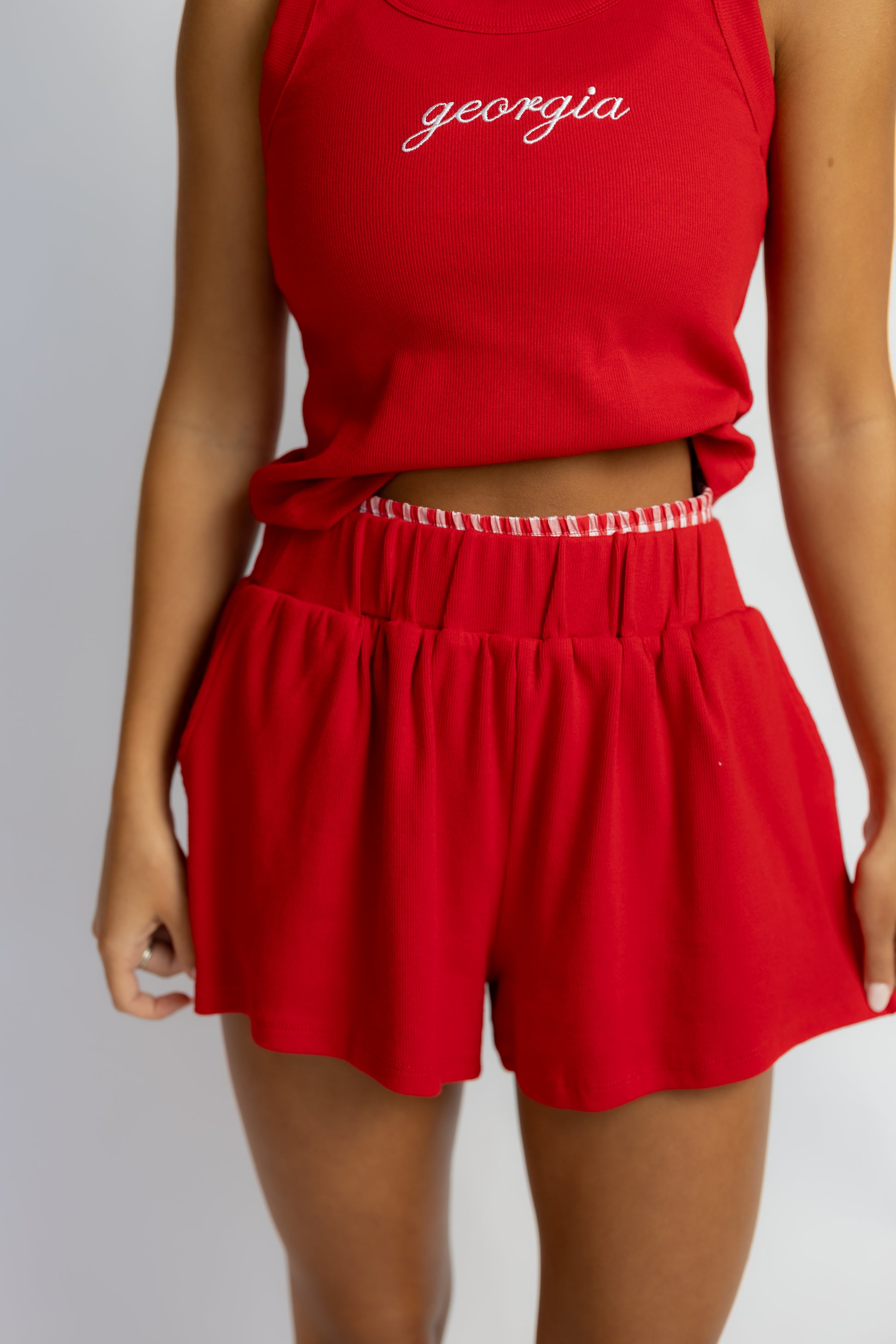 A model wearing a red sleeveless crop top with 'georgia' embroidered in white script and matching red pleated shorts with a white gingham ruffled waistband, shown against a light gray background.