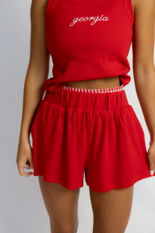 A model wearing a red sleeveless crop top with 'georgia' embroidered in white script and matching red pleated shorts with a white gingham ruffled waistband, shown against a light gray background.