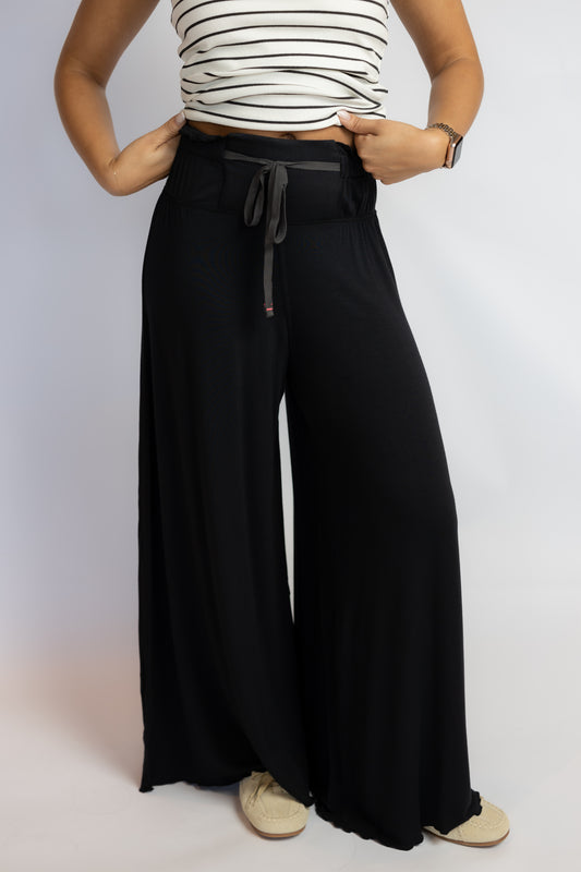 A person wearing black wide-leg pants with a dark drawstring waist, paired with a white and black striped top and beige shoes, shown against a plain light gray studio background.