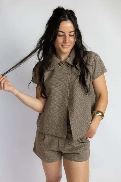 A woman modeling a matching brown tweed short-sleeve button-up shirt and shorts set against a plain light gray studio background.