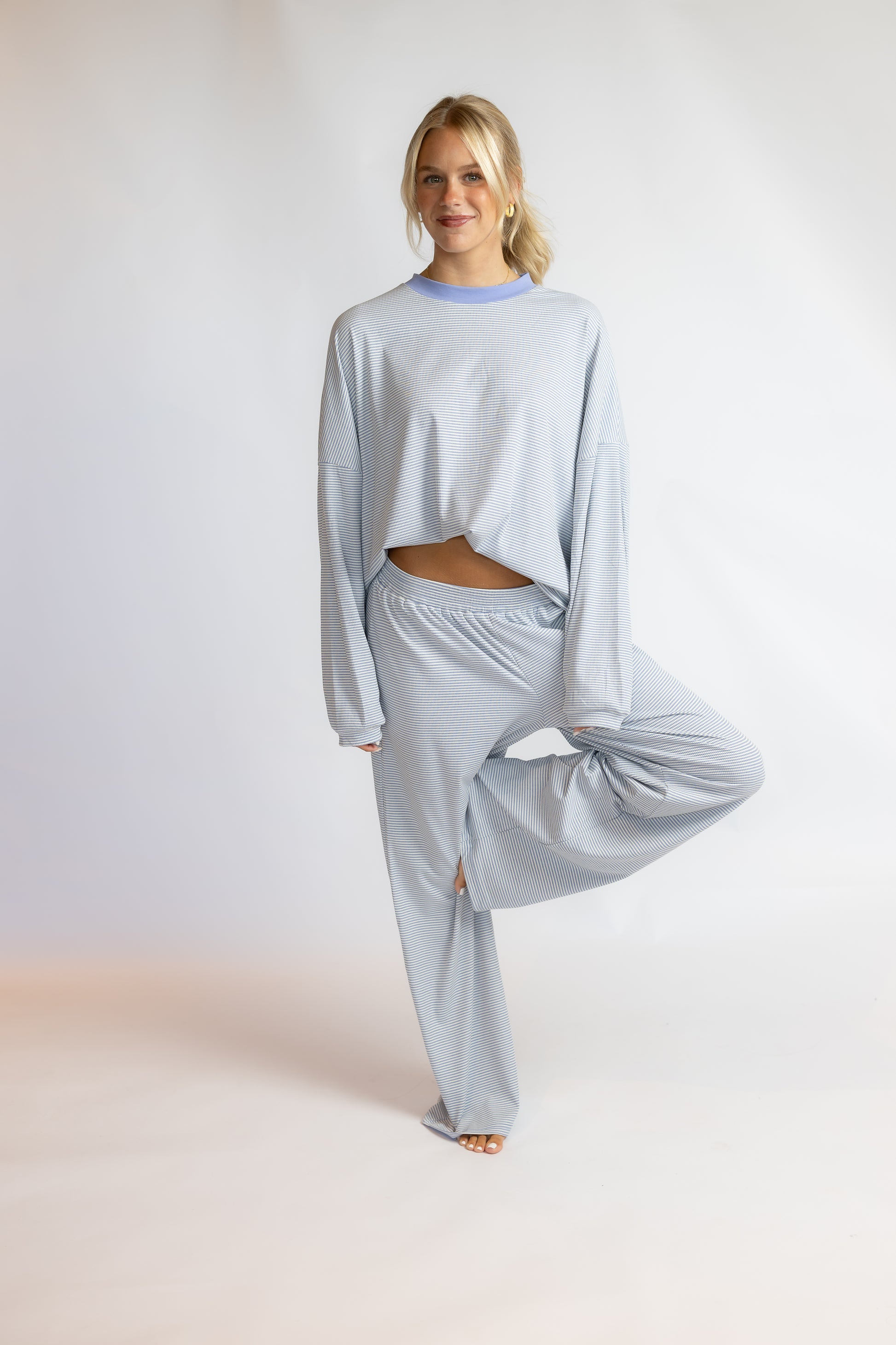 A woman modeling a light blue ribbed lounge set consisting of a cropped long-sleeve top and matching wide-leg pants, standing against a plain light gray studio background.