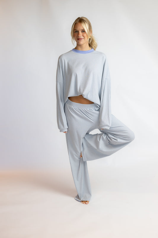 A woman modeling a light blue ribbed lounge set consisting of a cropped long-sleeve top and matching wide-leg pants, standing against a plain light gray studio background.