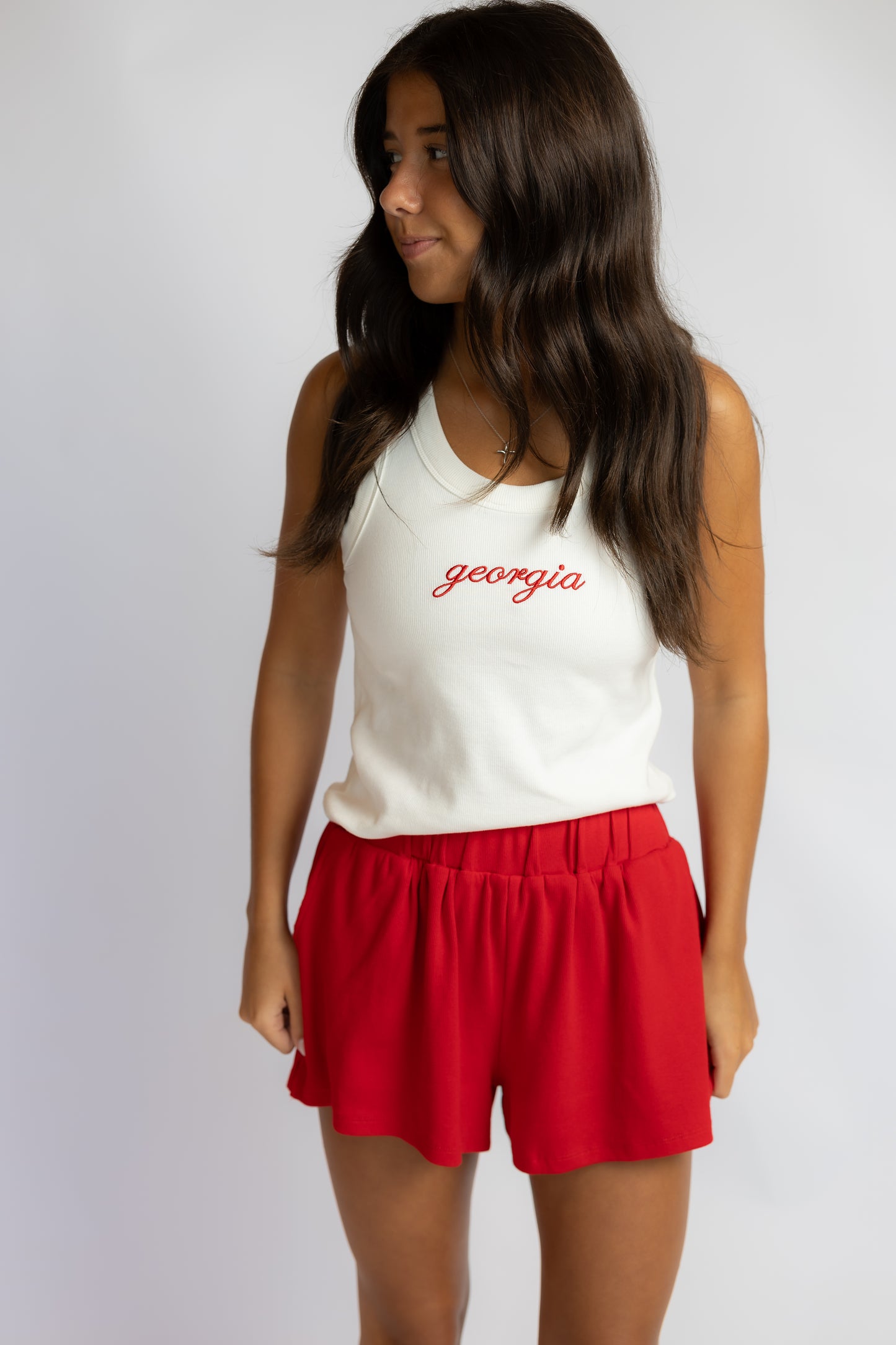 A woman modeling a white sleeveless tank top with 'georgia' embroidered in red script across the chest, paired with red shorts, against a light gray studio background.