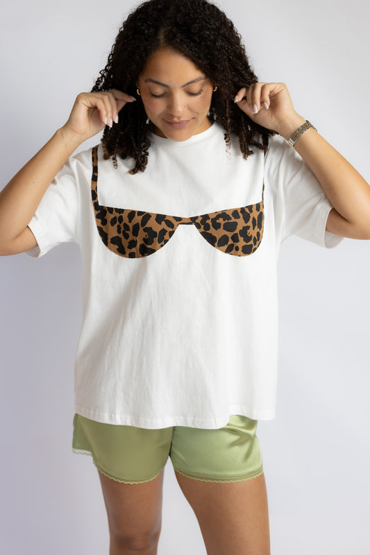 A woman wearing a white oversized t-shirt with a leopard-print bra graphic on the chest, paired with sage green shorts, against a light gray studio background.