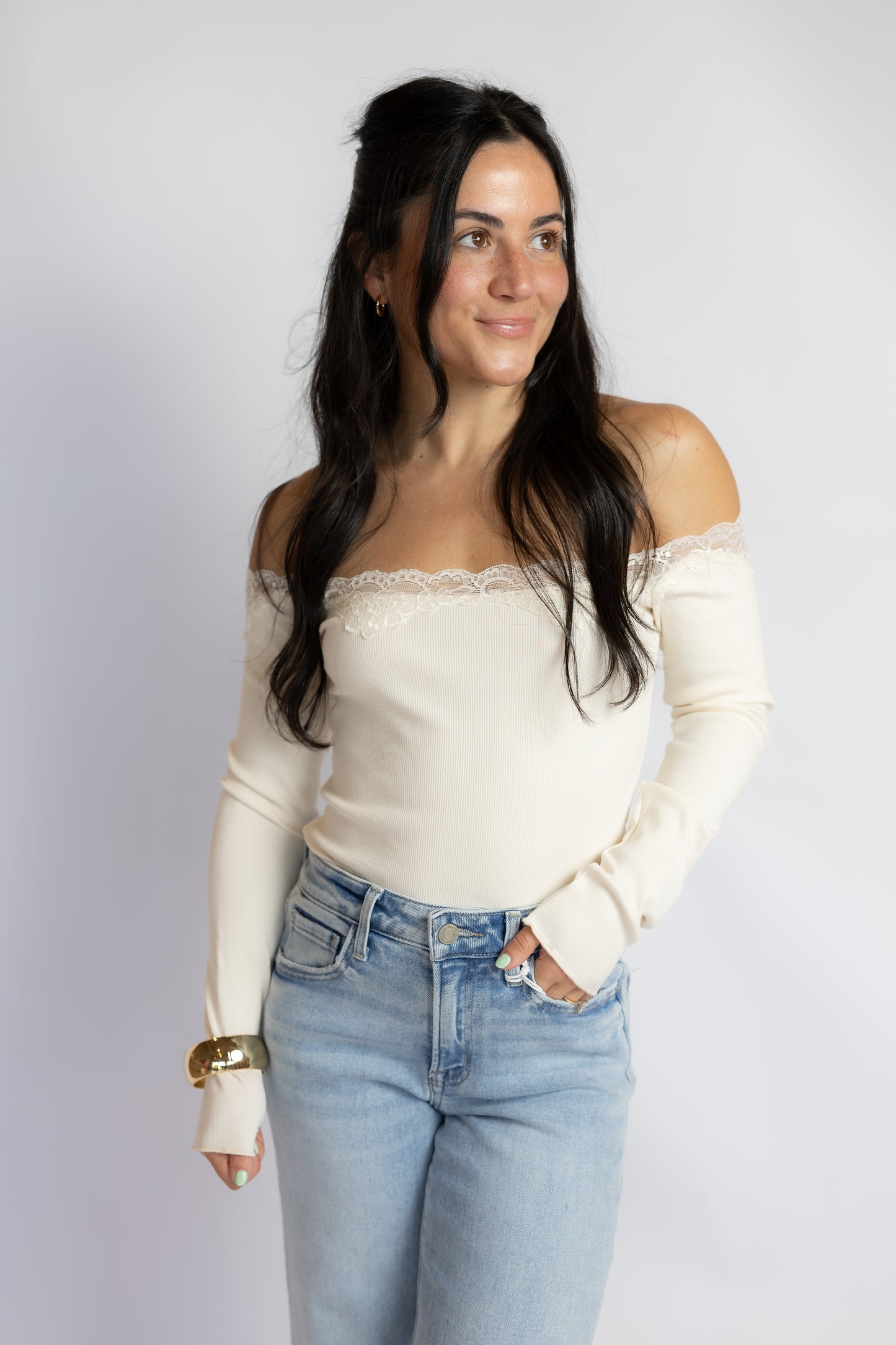 A woman modeling an off-the-shoulder cream ribbed knit top with a lace trim at the neckline, paired with light-wash blue jeans, against a plain light gray studio background.