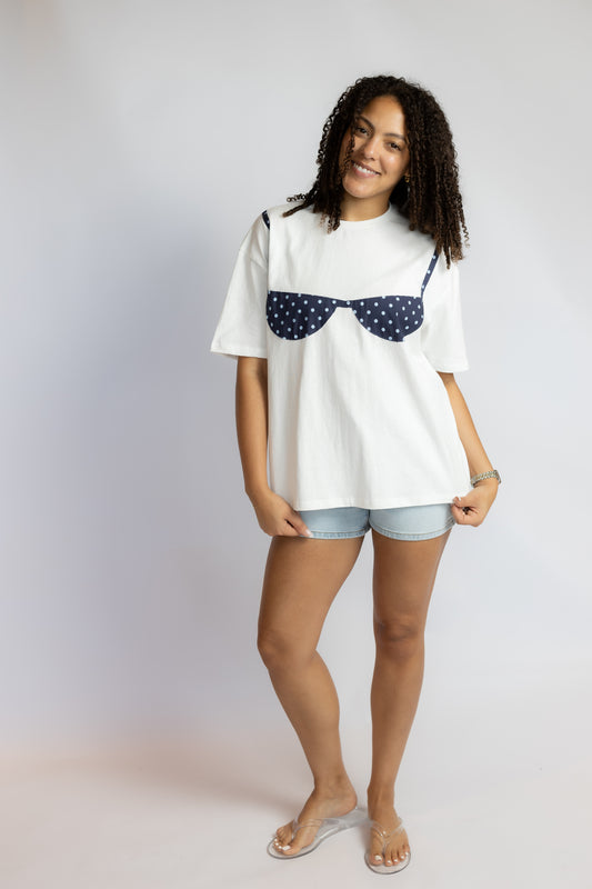 A woman modeling a white oversized t-shirt with a navy blue polka-dot bra graphic on the chest, paired with light denim shorts and clear flip-flops, against a light gray studio background.