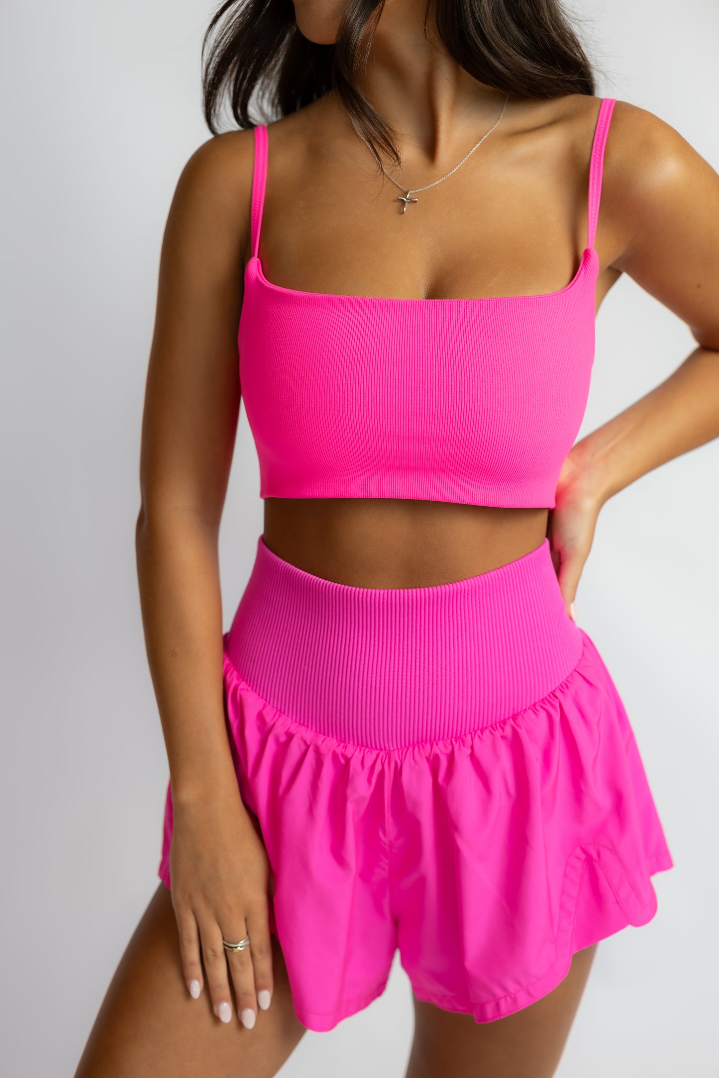 A woman modeling a bright neon pink two-piece activewear set consisting of a square-neck crop top with thin straps and high-waisted ruffled shorts, set against a plain light gray background.