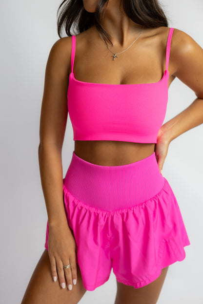 A woman modeling a bright neon pink two-piece activewear set consisting of a square-neck crop top with thin straps and high-waisted ruffled shorts, set against a plain light gray background.