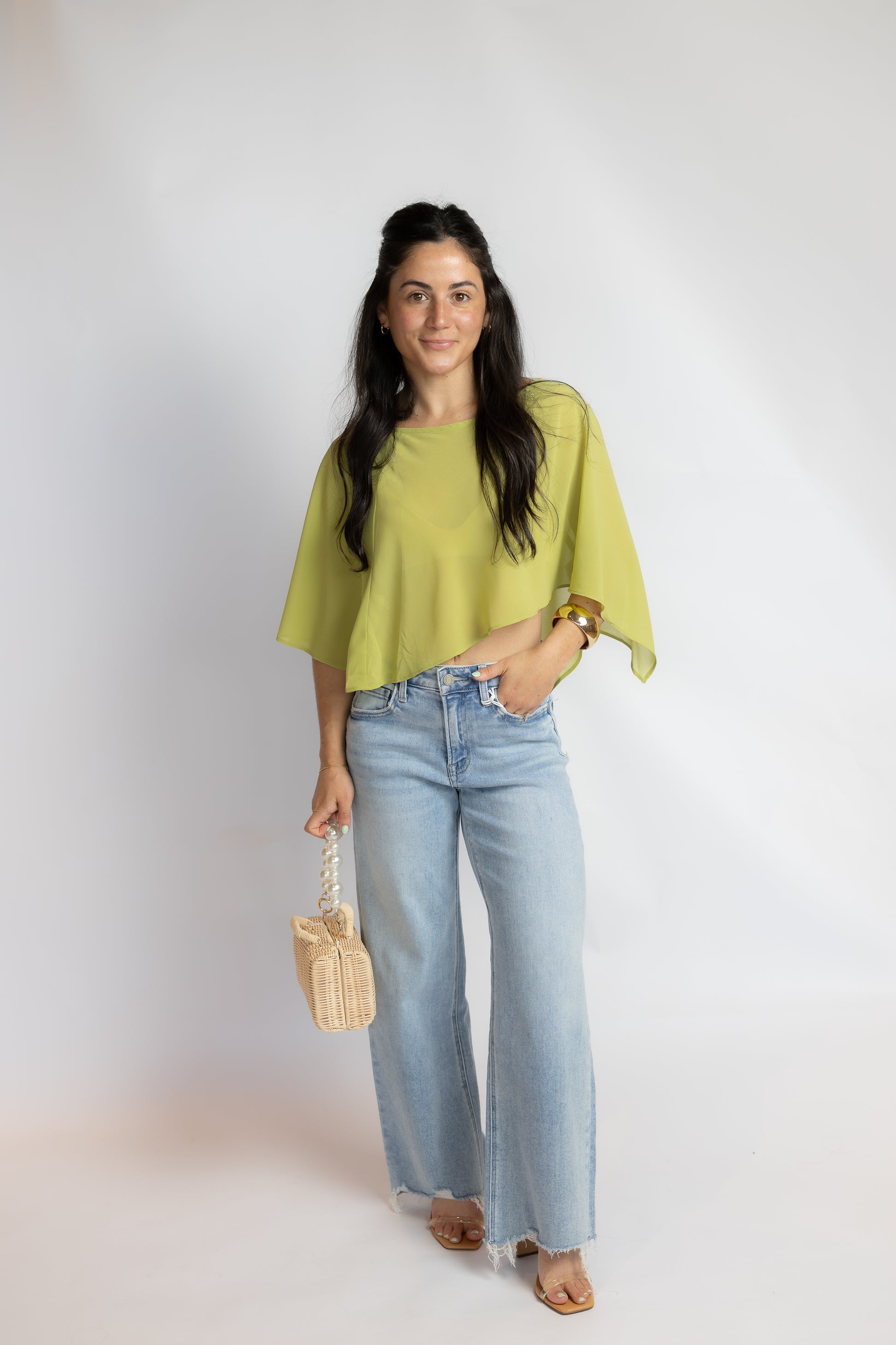 A woman modeling a light lime green short-sleeve top with wide, draped sleeves, paired with light-wash wide-leg jeans, holding a small woven straw bag against a plain white studio background.