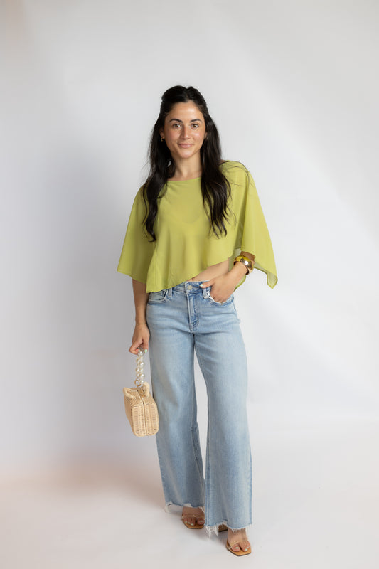 A woman modeling a light lime green short-sleeve top with wide, draped sleeves, paired with light-wash wide-leg jeans, holding a small woven straw bag against a plain white studio background.