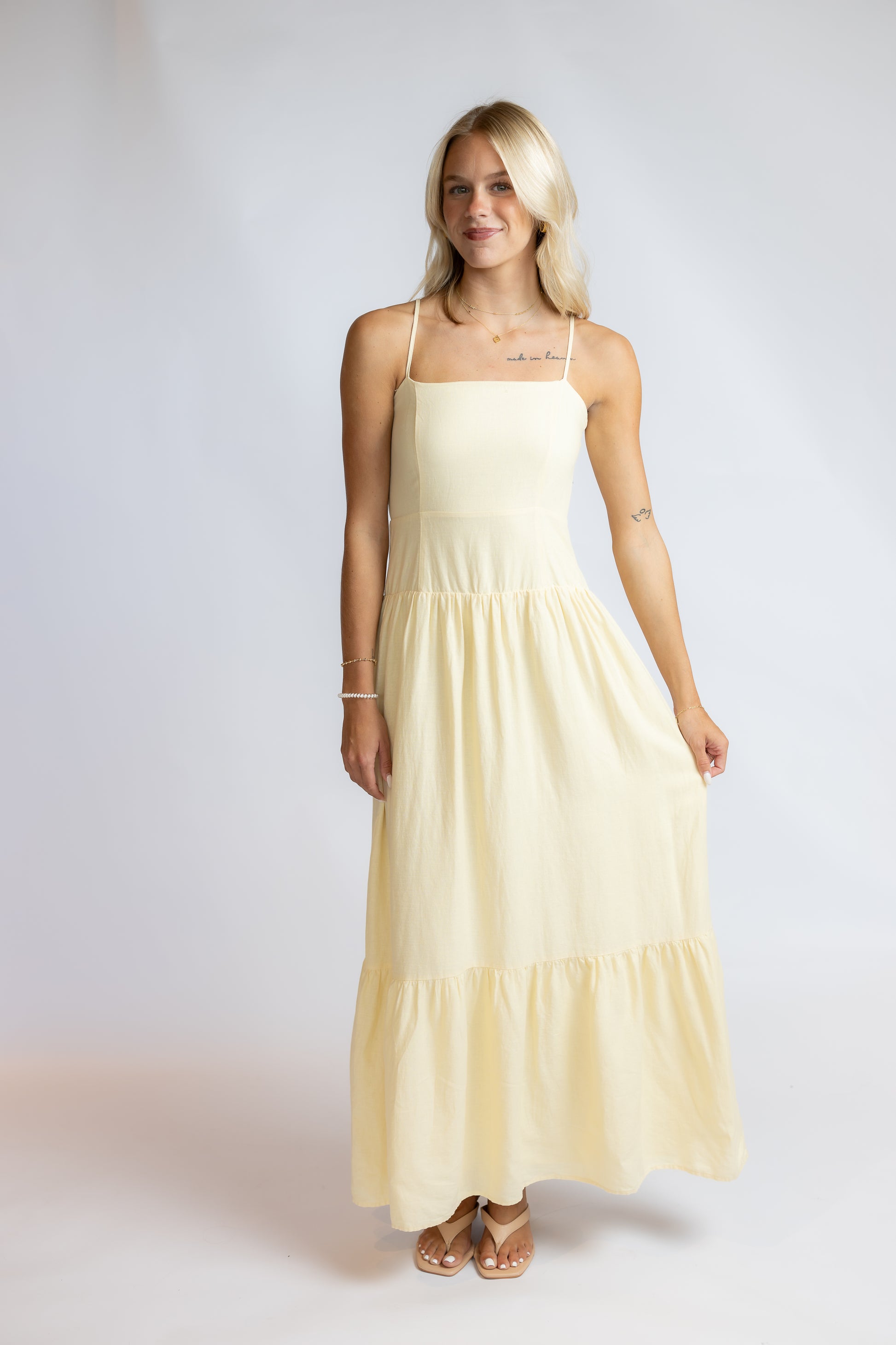 A blonde woman modeling a pale yellow maxi dress with thin spaghetti straps, a square neckline, and a tiered skirt, standing against a light gray studio background.