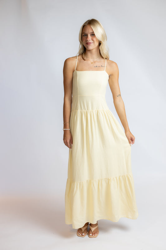 A blonde woman modeling a pale yellow maxi dress with thin spaghetti straps, a square neckline, and a tiered skirt, standing against a light gray studio background.