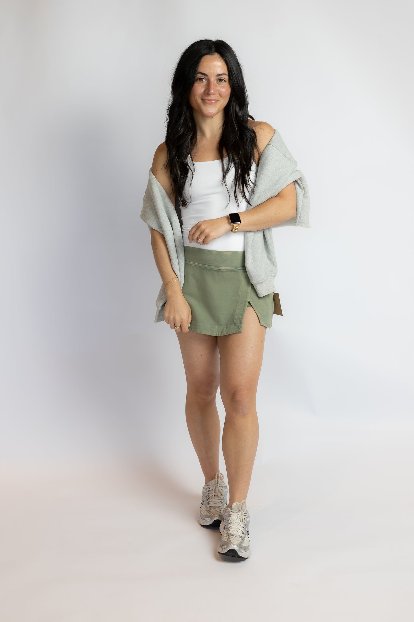 A woman modeling an olive green athletic mini skirt with side slits, paired with a white tank top, a light gray off-the-shoulder cardigan, and gray sneakers, against a plain light gray studio background.