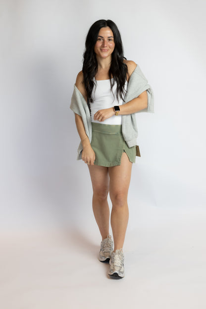 A woman modeling an olive green athletic mini skirt with side slits, paired with a white tank top, a light gray off-the-shoulder cardigan, and gray sneakers, against a plain light gray studio background.