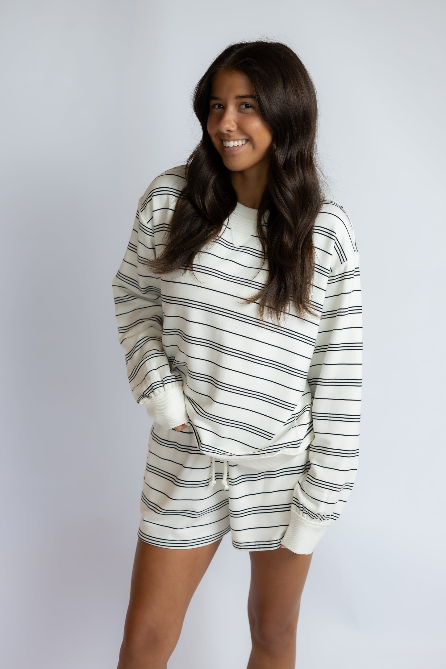 A woman modeling a matching two-piece loungewear set consisting of a long-sleeve sweatshirt and shorts, both featuring a horizontal navy blue stripe pattern on an off-white background, set against a plain light gray studio backdrop.