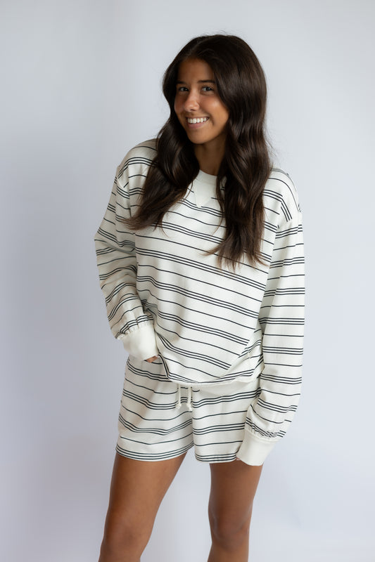 A woman modeling a matching two-piece loungewear set consisting of a long-sleeve sweatshirt and shorts, both featuring a horizontal navy blue stripe pattern on an off-white background, set against a plain light gray studio backdrop.