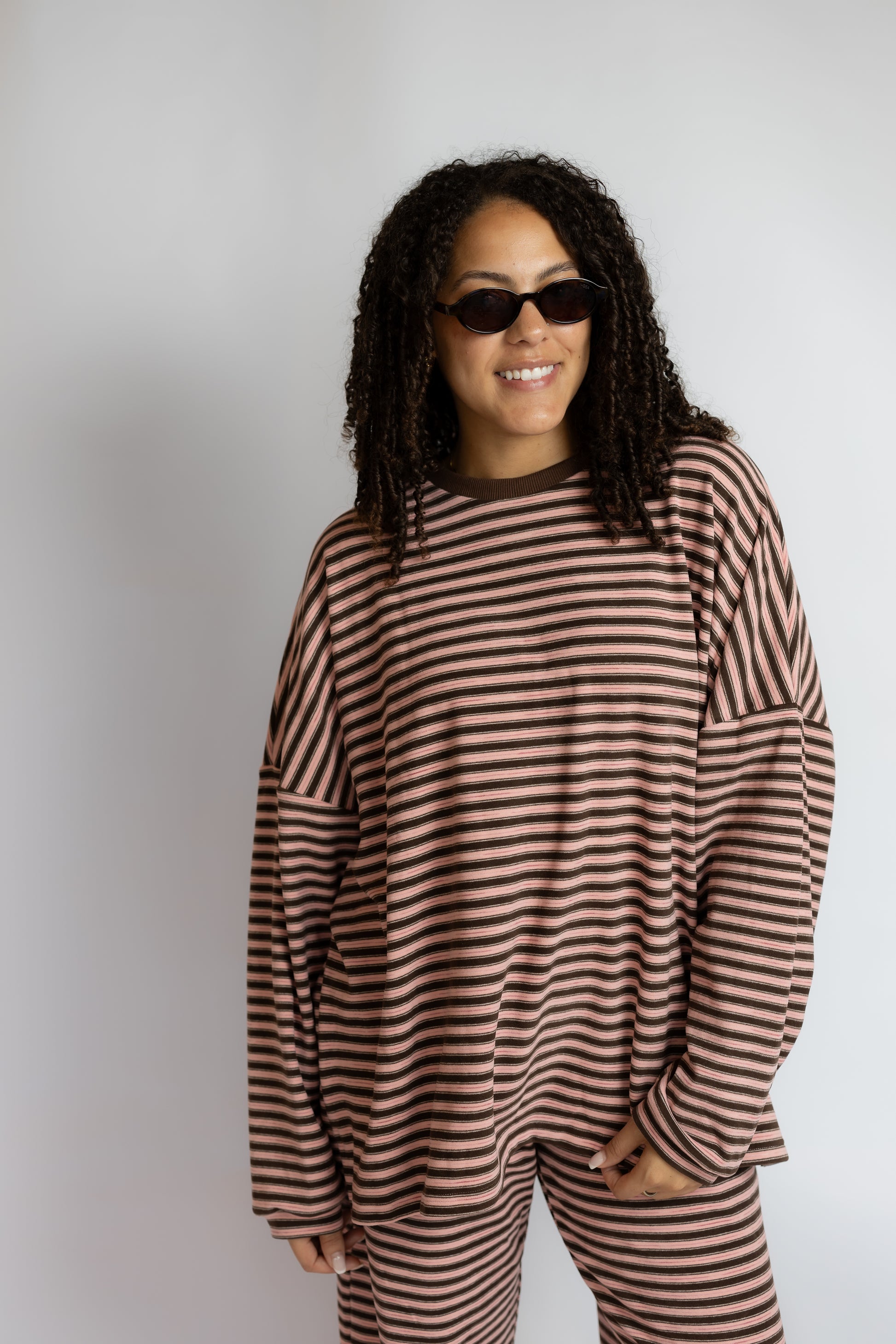 A woman with curly hair and sunglasses wearing a brown and beige striped long-sleeve top and matching striped pants, standing against a light gray studio background.