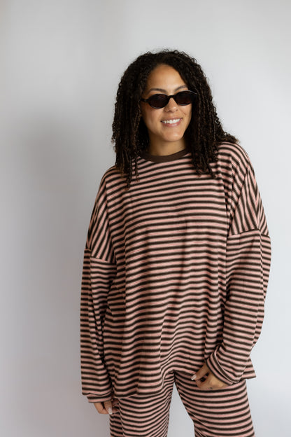 A woman with curly hair and sunglasses wearing a brown and beige striped long-sleeve top and matching striped pants, standing against a light gray studio background.