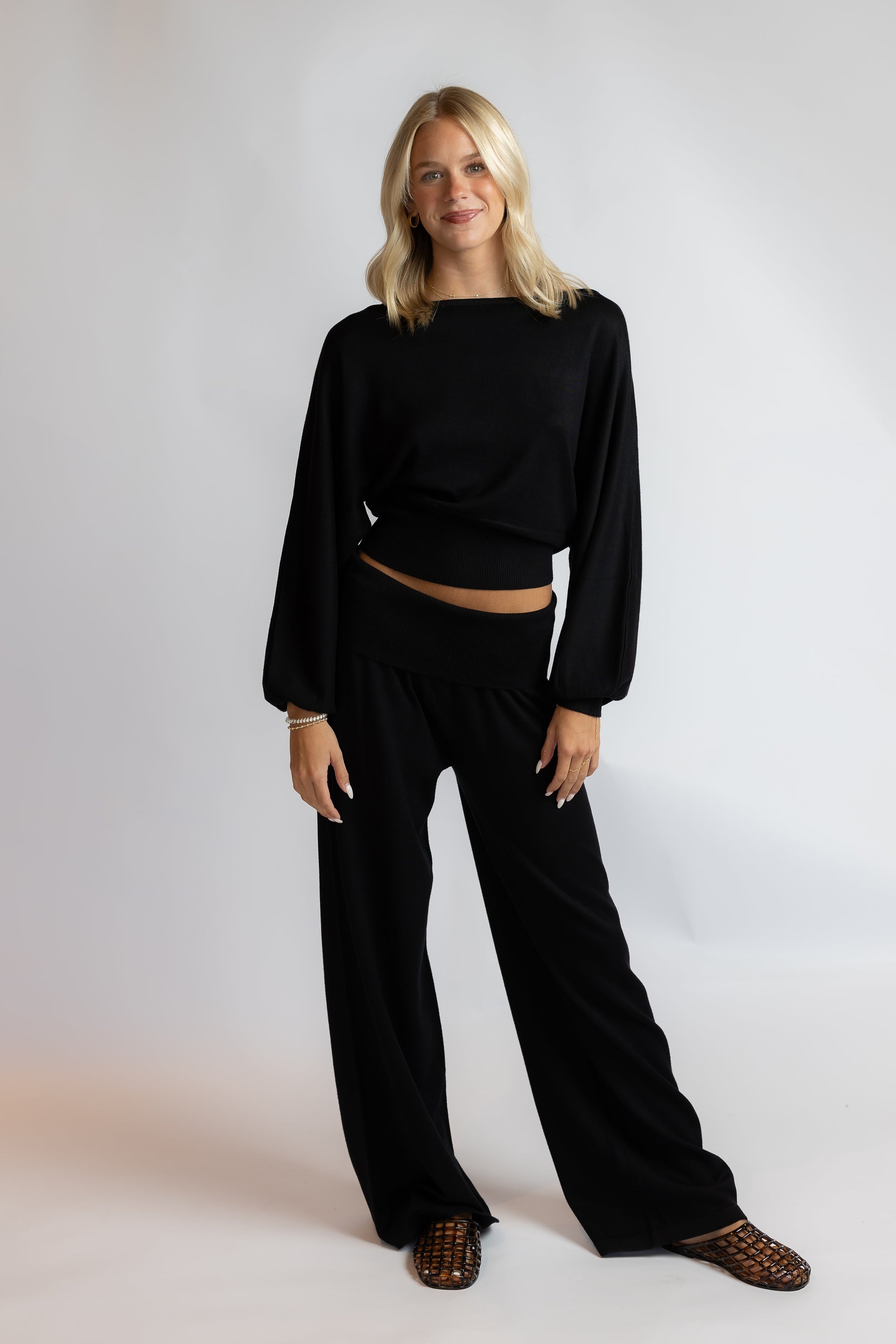 A woman modeling a black two-piece lounge set consisting of a cropped long-sleeve top and matching wide-leg pants, paired with brown woven flat shoes, against a light gray studio background.