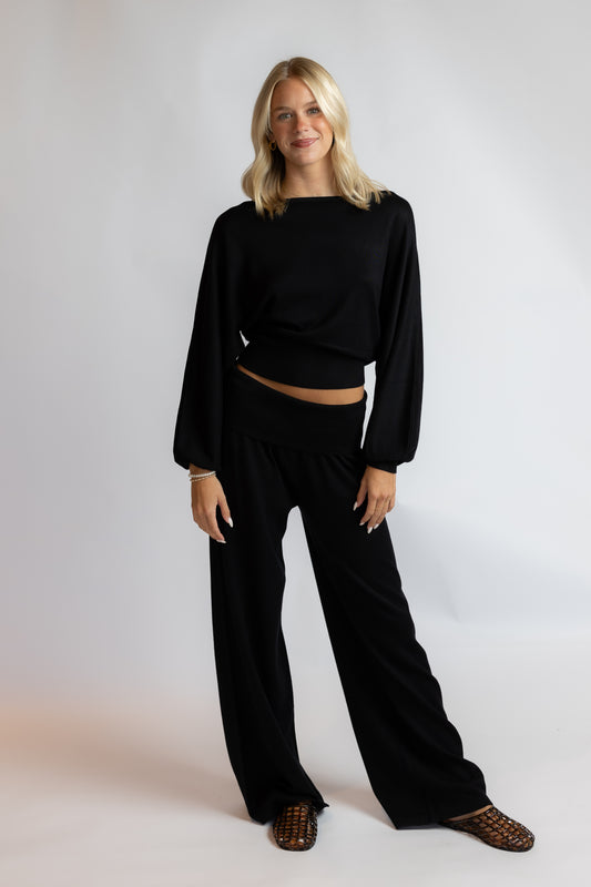 A woman modeling a black two-piece lounge set consisting of a cropped long-sleeve top and matching wide-leg pants, paired with brown woven flat shoes, against a light gray studio background.