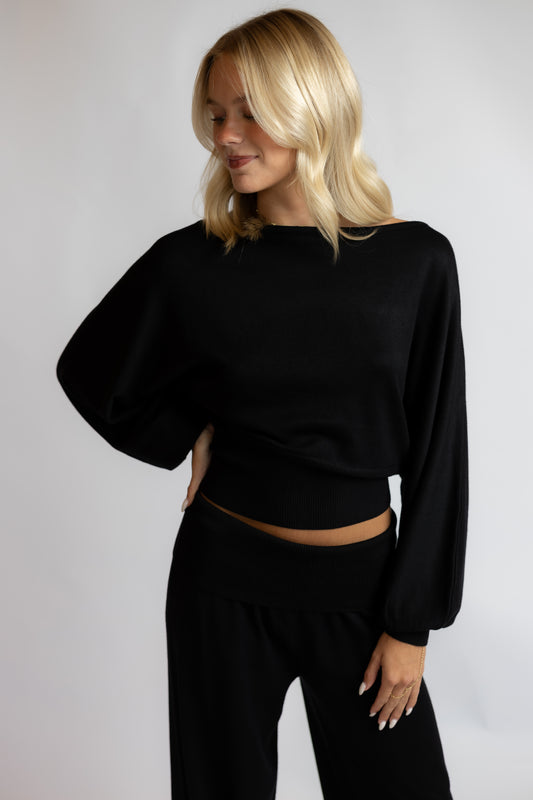 A blonde woman wearing a black long-sleeve cropped top and black pants, posing against a light gray studio background.