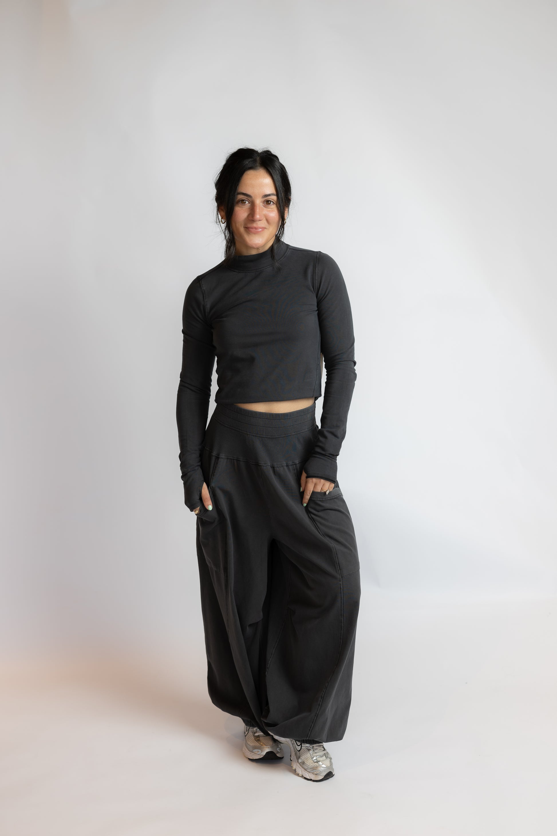 A woman modeling a black long-sleeve crop top and matching wide-leg pants, standing against a plain light gray studio background.
