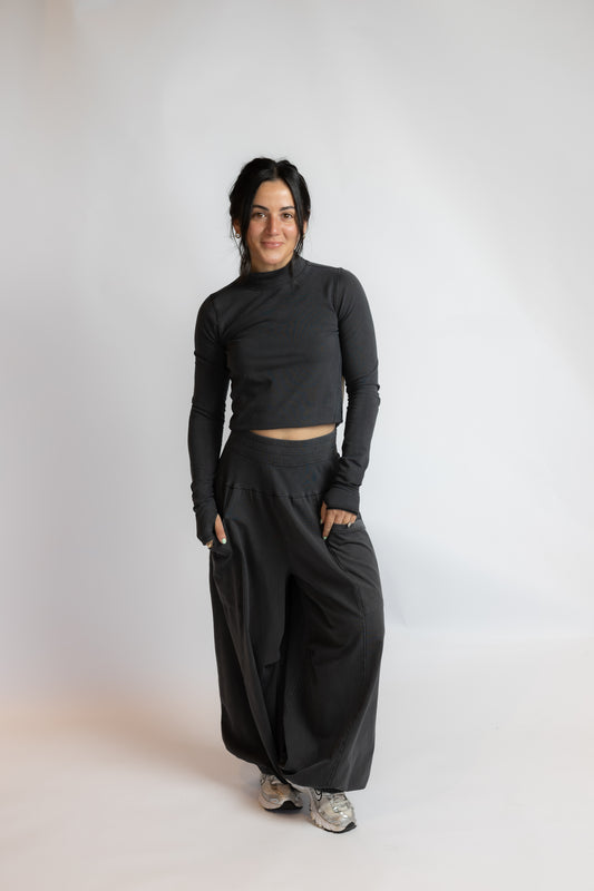 A woman modeling a black long-sleeve crop top and matching wide-leg pants, standing against a plain light gray studio background.