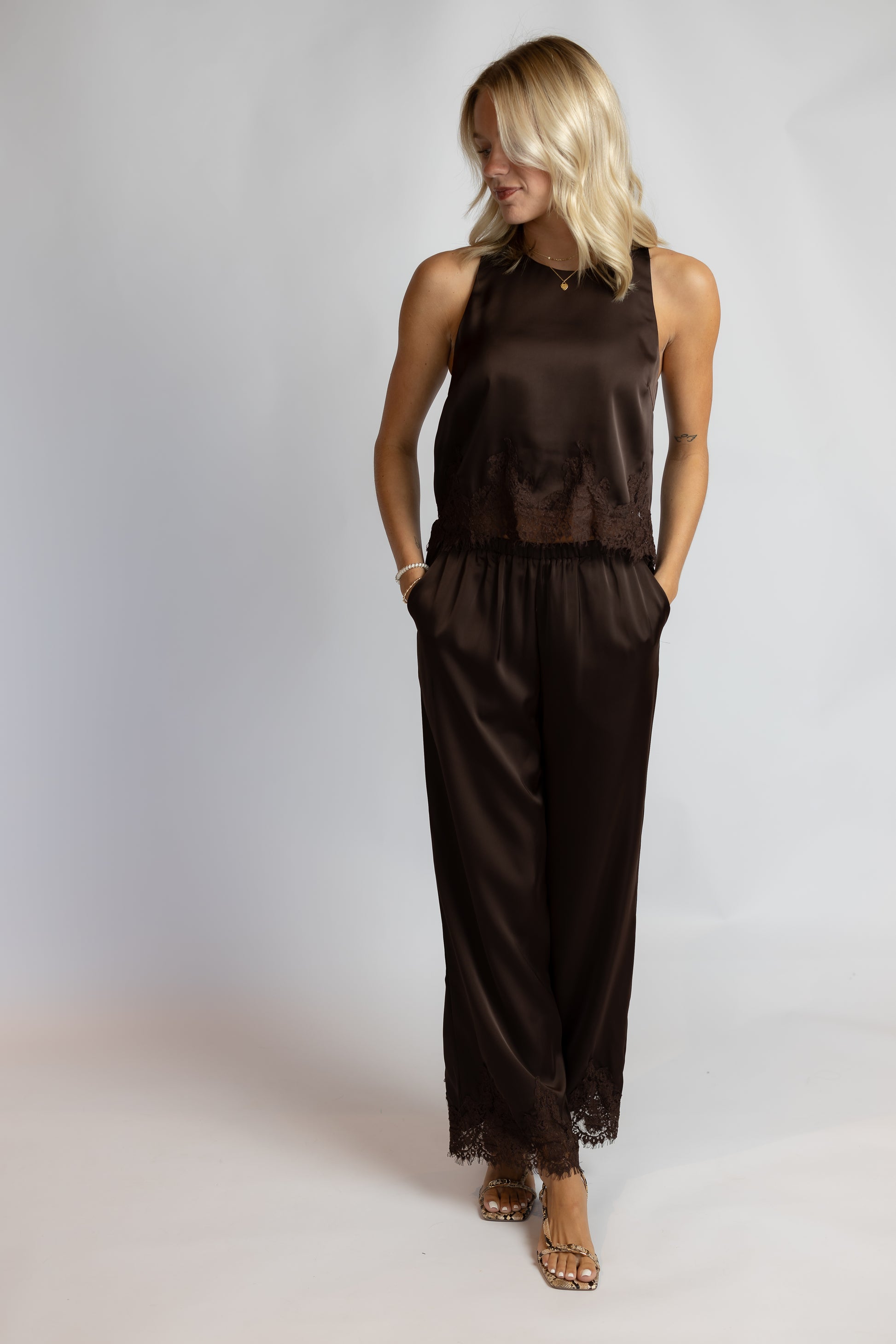 A woman modeling a dark brown satin jumpsuit with lace trim, standing against a light gray studio background.