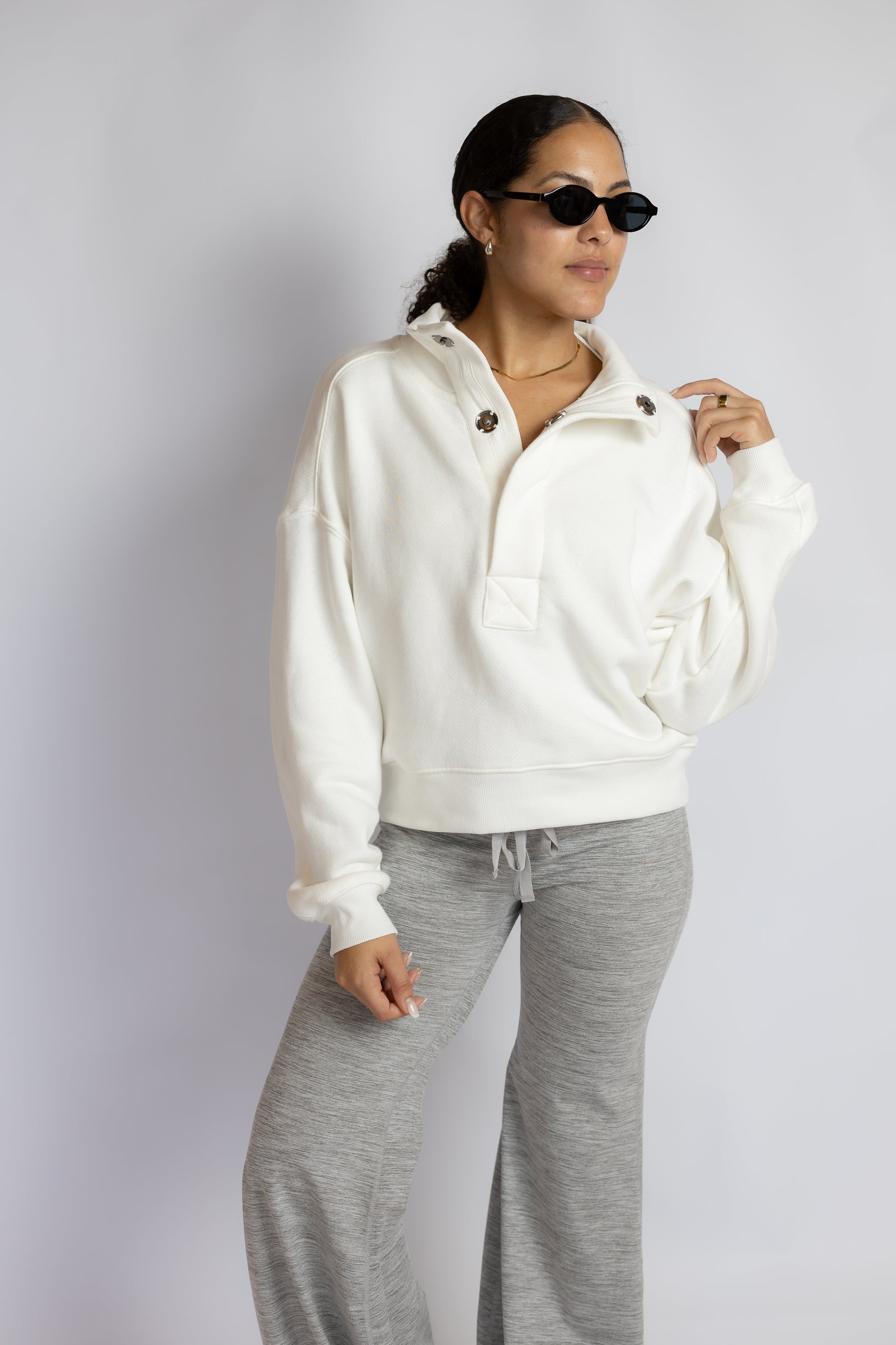 A woman modeling a white half-zip pullover hoodie with gray sweatpants, sunglasses, and a gold necklace against a light gray studio background.