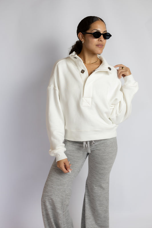A woman modeling a white half-zip pullover hoodie with gray sweatpants, sunglasses, and a gold necklace against a light gray studio background.